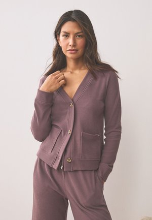 Kvinde med langt mørkt hår iført en mauve ribbet cardigan og matchende bukser, stående mod en ensfarvet lys baggrund med den ene hånd nær kraven.