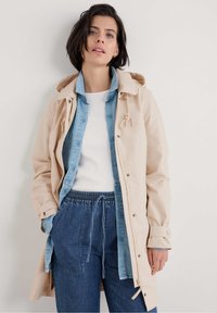 Veste à capuche beige avec cordon de serrage, superpos�ée sur une chemise en denim bleu clair, un haut blanc côtelé et un pantalon en denim avec taille élastique.
