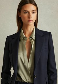 Kvinde med lige brunt hår iført en mørk tekstureret blazer over en olivengrøn satinbluse, der kigger let til siden mod en ensfarvet baggrund.