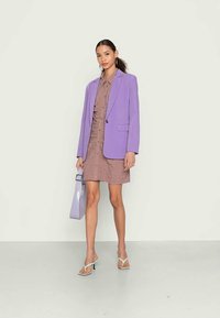Blazer lavanda, vestido em botão castanho e mala lavanda. O modelo veste sandálias de salto verdes. Texturas suaves e design moderno.