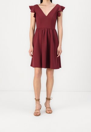 Nainen, jolla on polvipituinen viininpunainen mekko, jossa liehuvat hihat ja V-aukko, yhdistettynä vaaleanruskeisiin remmikenkiin.