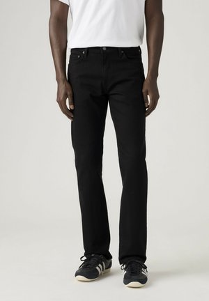 Man draagt zwarte jeans met rechte pijpen, wit T-shirt en zwarte sneakers met witte strepen, staand tegen een effen achtergrond.