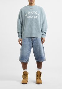 Lichtblauwe geribde trui met witte tekst, gecombineerd met blauwe denim bermuda shorts en tan laarzen met gele veters. Witte gemusterde sokken zijn zichtbaar.