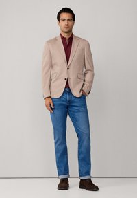 Beiger Blazer mit taillierter Passform, zwei vorderen Taschen, schwarzen Knöpfen, kombiniert mit einem bordeauxroten Poloshirt und blauen Jeans, getragen mit braunen Stiefeln.