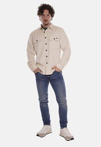Camicia in corduroy beige con due tasche sul petto, bottoni neri, indossata sopra una camicia verde scuro. Abbinata a jeans blu e scarpe da ginnastica bianche.