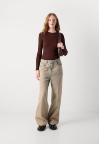 Hnědá dlouhá halenka s dlouhým rukávem, volné modré denimové džíny a černé boty. Modelka drží hnědou kabelku. Hladká textura, uvolněný střih, viditelné detaily šití.