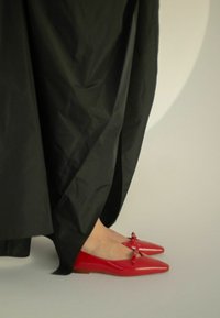 Ballerine in vernice rossa con punta affusolata e piccoli accenti a fiocco, indossate con un lungo capo nero, che mette in risalto una texture liscia e lucida.