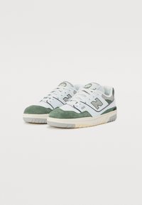 Baltos sportbačiai su žaliais zomšos akcentais, guminėmis padais, tekstūruotu audiniu, apvaliu priekiu ir siuvinėtu logotipu ant liežuvėlio. Dizainas yra žemas ir klasikinis.