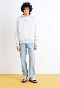 Hoodie gris clair avec une poche frontale et du texte vert, associé à un jean bleu clair et des baskets multicolores. Design minimaliste.