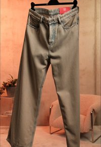 Beige straight-leg jeans hængende på en sort bøjle med rød størrelsesmærkat, sat op mod et varmt tonerum med en blød stol og en potteplante i baggrunden.