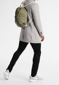 Une personne portant un manteau gris, un pantalon noir et des baskets grises marche avec un sac à dos à cordon de couleur vert olive.