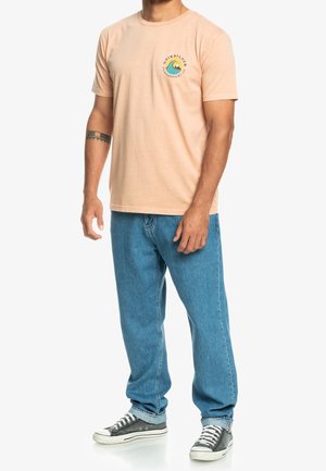 Uomo che indossa una T-shirt grafica color pesca chiaro, jeans blu, sneaker bianche e nere, in piedi con una postura rilassata su uno sfondo bianco.