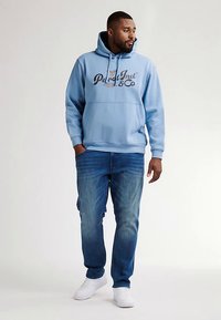 Modrá bavlněná mikina s kapucí a přední kapsou s grafickým logem, ve volném střihu, doplněná světle modrými džínami a bílými teniskami.