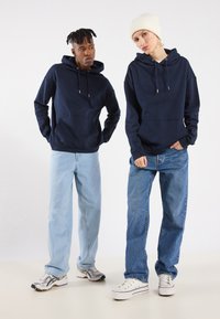 Marinegenser med frontlommer og snor; lyseblå og mørkeblå jeans; en i en hvit lue, den andre i joggesko.