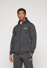 Šedý zipsový hoodie s kapucňou, s malým logom. Vyrobený z mäkkého materiálu, má postranné vrecká a rebrované manžety. Viditeľné sú aj zodpovedajúce tepláky.