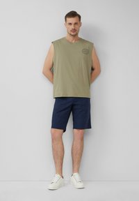 Maglietta senza maniche verde oliva con collo rotondo e logo, abbinata a pantaloni corti blu navy. Il modello indossa sneakers bianche, in piedi davanti a uno sfondo neutro.