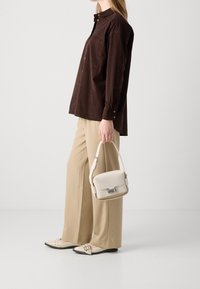 Camicia button-up marrone con maniche lunghe, pantaloni a gamba larga crema, che tiene una borsa crema con accessori in argento e scarpe flat crema.