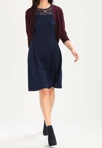 Kvinna som går i en marinblå klänning med spetskrage, burgundy kortärmad kofta och svarta ankelstövlar mot vit bakgrund.