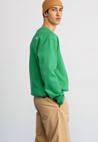 Grønn sweatshirt med rippekanter på ermene og halsen, med en subtil logo på baksiden. Kombinert med beige bukser, satt mot en ensfarget bakgrunn.