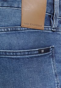 Denim jeans met een bruine leren taillebandlabel met de tekst "NO EXCESS" en een klein blauw accentlabel op de achterzak. Lichtblauwe wassing.