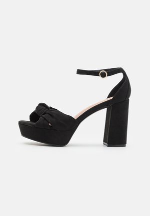 Sort ruskind platform sandal med chunky høj hæl, knude foran rem og justerbar ankelrem med guldlås.