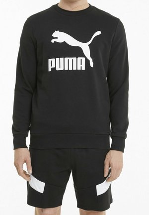 Czarna bluza z długimi rękawami, ściągaczami na rękawach i dole. Na przodzie znajduje się biały logo Puma oraz napis. Do tego czarne spodenki z białymi akcentami.