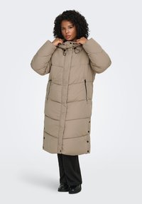 Lång, beige vadderad jacka med hög krage, frontdragkedjor och knappar vid fållen. Har ett quiltat mönster och sidofickor.