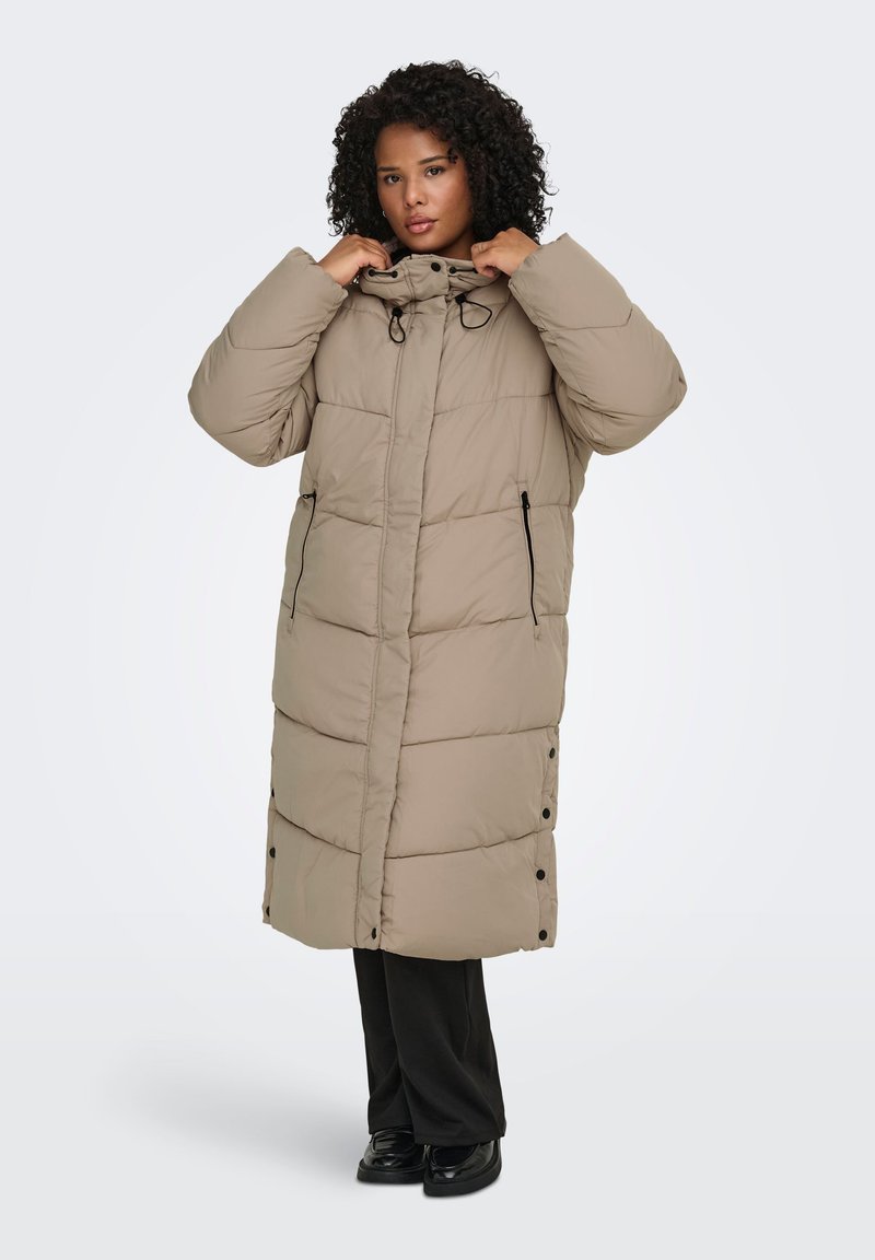 Lång, beige vadderad jacka med hög krage, frontdragkedjor och knappar vid fållen. Har ett quiltat mönster och sidofickor.