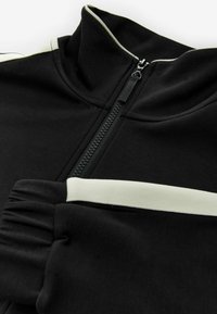 Veste zippée noire avec un col montant, en tissu texturé, et une bande blanche contrastante le long de l'épaule et des poignets.