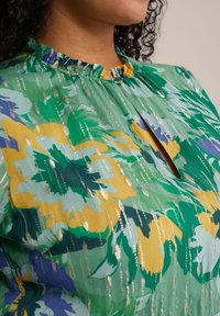 Blouse florale verte avec un col rufflé. Présente des teintes jaunes, bleues et vertes, avec des accents métalliques et un petit détail en forme de trou de serrure.