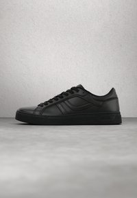Baskets basses en cuir noir avec lacets et logo discret de la marque sur le côté et la semelle, posées sur une surface texturée grise.