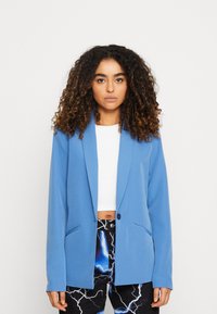 Blazer azul con cierre de un solo botón, corte ajustado, solapas tipo muesca y dos bolsillos laterales, llevado sobre una blusa blanca y pantalones con estampado.