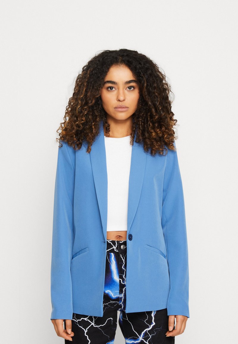 Blazer azul con cierre de un solo botón, corte ajustado, solapas tipo muesca y dos bolsillos laterales, llevado sobre una blusa blanca y pantalones con estampado.