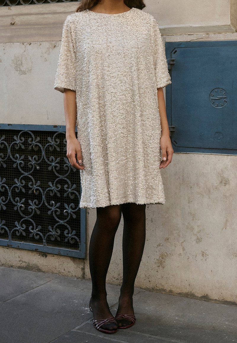 Personne portant une robe texturée crème longueur genoux, des collants noirs et des talons ouverts, debout sur un trottoir près d'un mur de bâtiment.