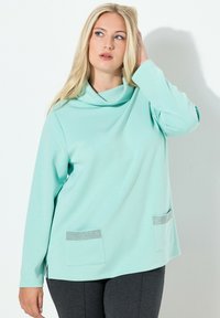 Długi top z długim rękawem w kolorze jasnej mięty z luźnym kołnierzem typu komin, z dwoma przednimi kieszeniami ozdobionymi dekoracyjnymi kryształkami. Miękki, gładki materiał.