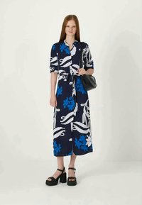 Vestido maxi com padrão floral, apresentando designs em branco e azul sobre um fundo azul-marinho, com cintura cingida e mangas arregaçadas. Combinado com sandálias plataforma pretas.