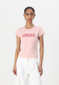 Šviesiai rožinė medvilninė marškinė su trumpomis rankovėmis, turi tekstūruotą tamsiai rožinę "GUESS" logotipą. Lygi medžiaga. Taisyklinga figūrai pritaikyta forma.