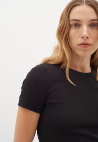 Svart ribbad t-shirt med rund halsringning och korta ärmar, tillverkad av ett mjukt, stretchigt tyg. Närbild av överkroppen.