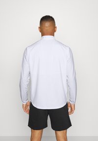 Camisa blanca de manga larga con un cuello alto, textura suave y dobladillo redondeado. Llevada con pantalones cortos negros, mostrando un ajuste relajado y un diseño minimalista.