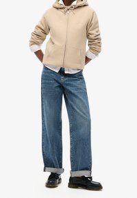 Sweatshirt à capuche beige zippé sur une chemise blanche, associé à un jean large bleu clair, avec des poignets retroussés, et des chaussures noires.