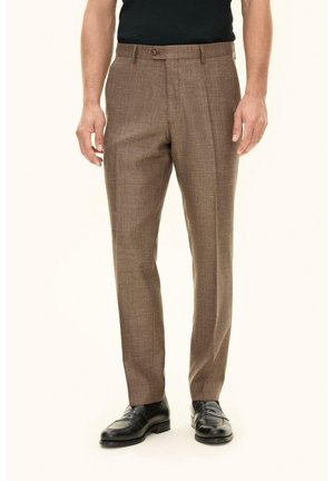 Mies, jolla on yllään ruskeat räätälöidyt housut, mustat nahkaiset loaferit ja musta hihaton paita, seisoo rentoina käsivarsin.