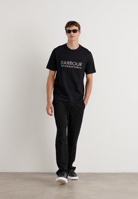 Camiseta preta com o logótipo "BARBOUR INTERNATIONAL", combinada com calças pretas e sapatos pretos. Modelo a usar óculos de sol escuros, de pé contra um fundo claro.
