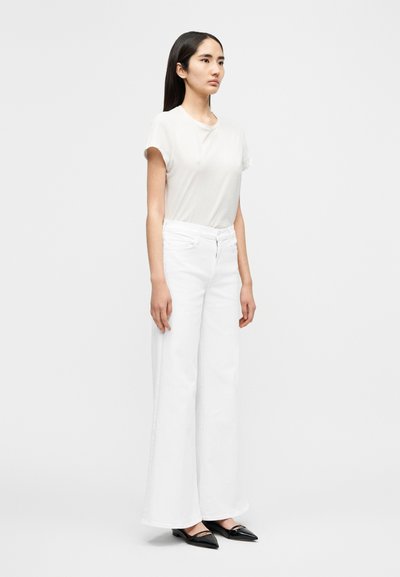 Άσπρη βαμβακερή μπλούζα με κοντά μανίκια, συνδυασμένη με άνετο λευκό τζιν και μαύρα μπαλαρίνες με μυτερή μύτη, εμφανίζεται από το πλάι.