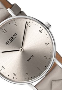 Montre en quartz argentée avec un cadran en métal brossé, des aiguilles noires et des marqueurs horaires numériques. Bracelet en cuir gris doux avec des accents texturés.