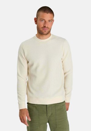 Mann trägt einen strukturierten cremefarbenen Langarmpullover und grüne Cargohosen, steht vor einem schlichten Hintergrund.