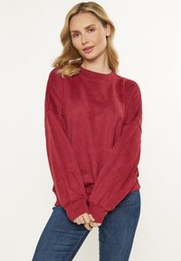 Burgundy színű pulóver hosszú ujjakkal, bordázott nyakkivágással és laza szabással. Kék farmernadrággal párosítva, amely puha textúrával és letisztult dizájnnal rendelkezik.