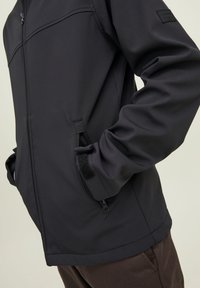 Veste softshell noire avec poches zippées et poignets réglables en Velcro portée par une personne avec les mains dans les poches, montrée du milieu du torse jusqu'aux hanches.