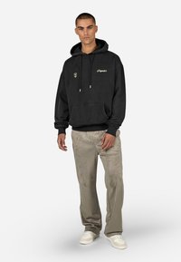 Čierna mikina s kapucňou a vreckom na prednej strane, s farebnými šnúrkami a vyšitým logom. Šedé, textúrované nohavice s širokými nohavicami a biele tenisky dotvárajú outfit.