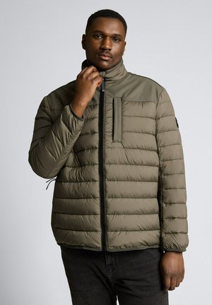 Man draagt olijfgroene doorgestikte jas met ritskraag, houdt rits bij de nek, staat tegen een effen grijze achtergrond.