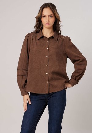 Femme aux cheveux châtain mi-longs, portant une chemise marron à manches longues boutonnée et un jean bleu foncé, debout avec une main sur la hanche.